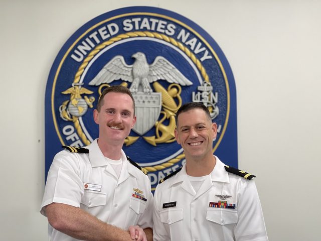 Two Navy officers in dress whites in front of the Navy seal